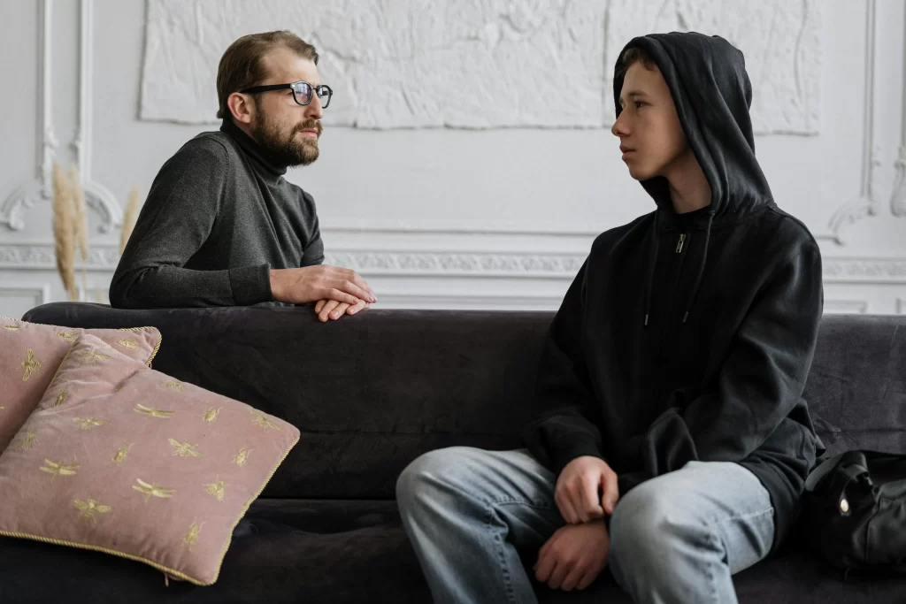 A therapist speaks with a teenage boy in a hoodie during a counseling session, seated on a couch in a calm, indoor setting.