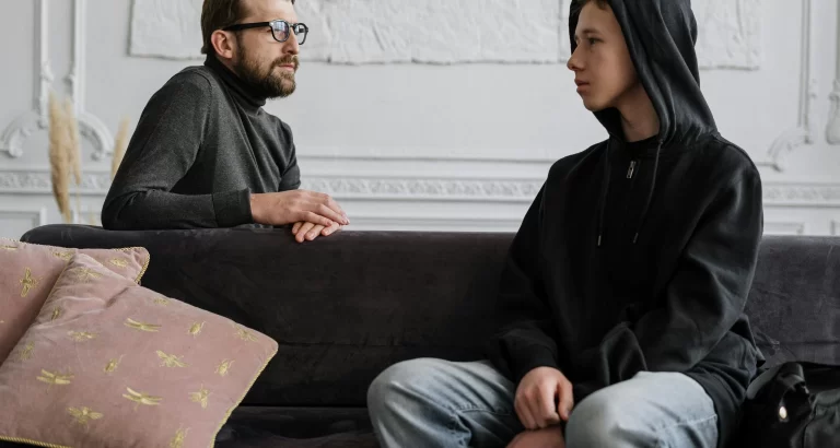 A therapist speaks with a teenage boy in a hoodie during a counseling session, seated on a couch in a calm, indoor setting.