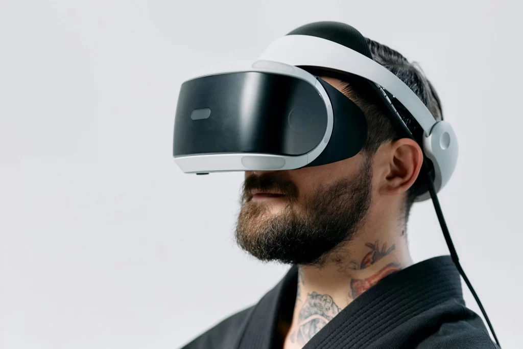 A man with a beard and neck tattoos wears a VR headset, immersed in a virtual reality experience against a plain background.