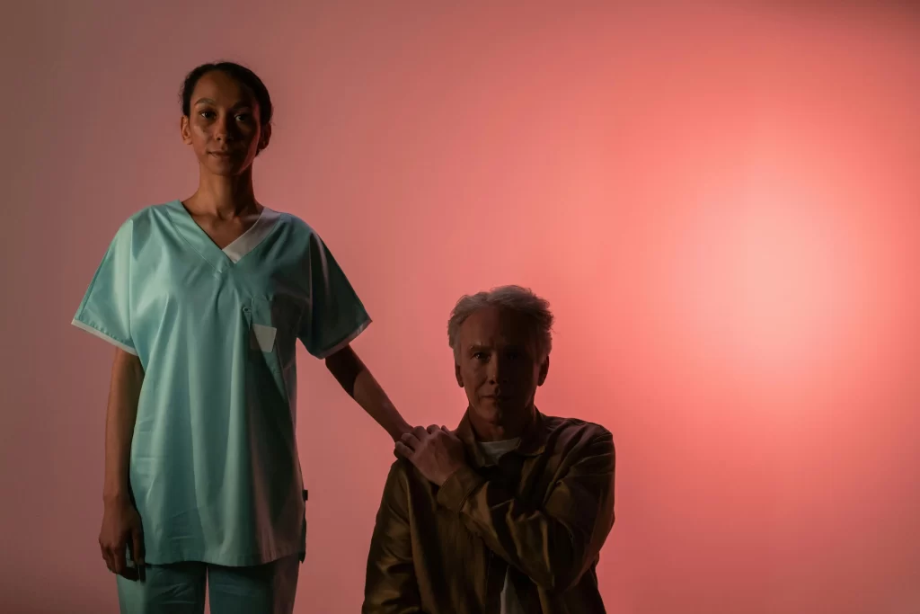 A healthcare worker stands beside an elderly man, resting a hand on his shoulder, against a warm pink background.