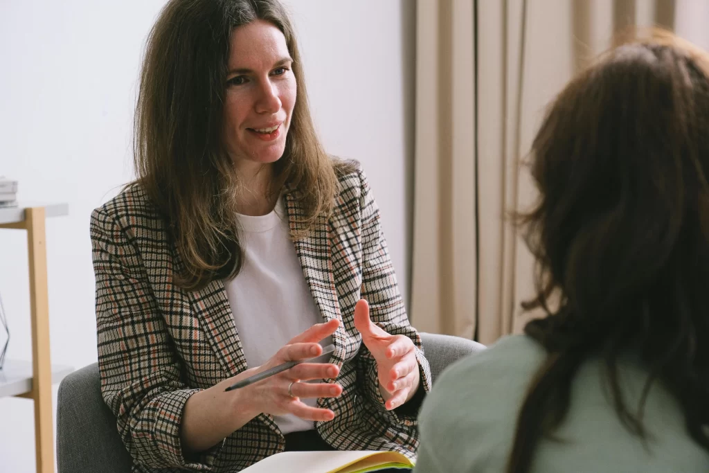 A healthcare professional engages in a one-on-one coaching session, speaking with a client in a relaxed indoor setting.