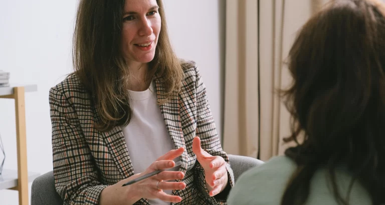 A healthcare professional engages in a one-on-one coaching session, speaking with a client in a relaxed indoor setting.